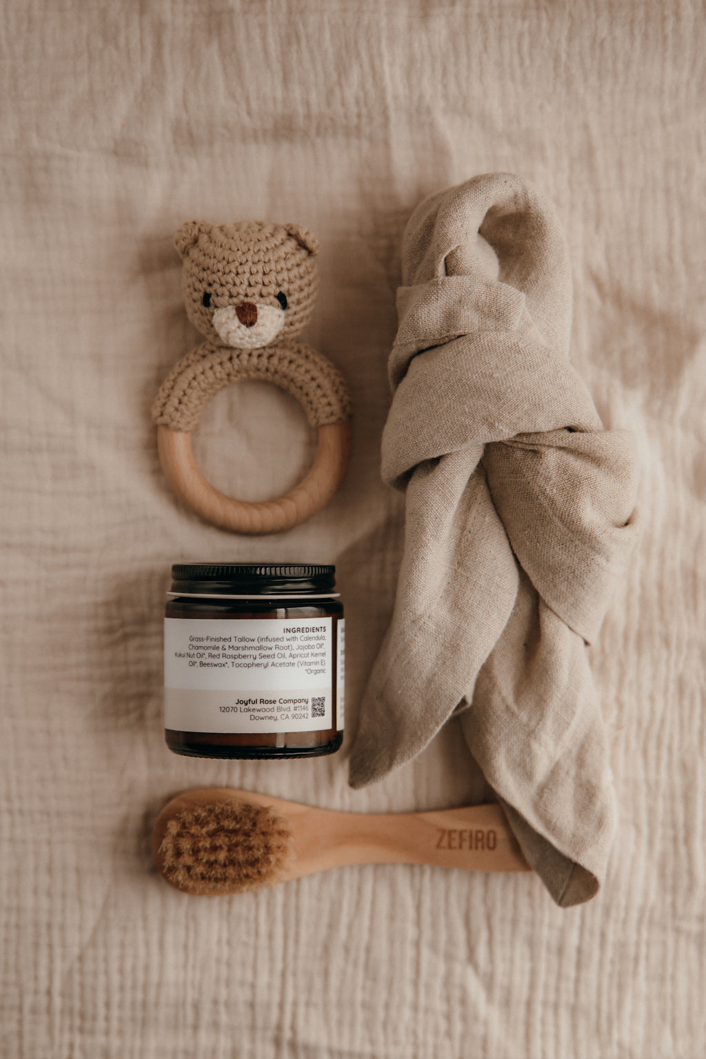 Baby care items including a teething ring, jar, towel, and brush on a textured surface.
