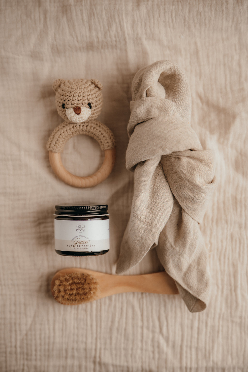 Baby care items including a wooden ring, jar, towel, and brush on a soft surface.