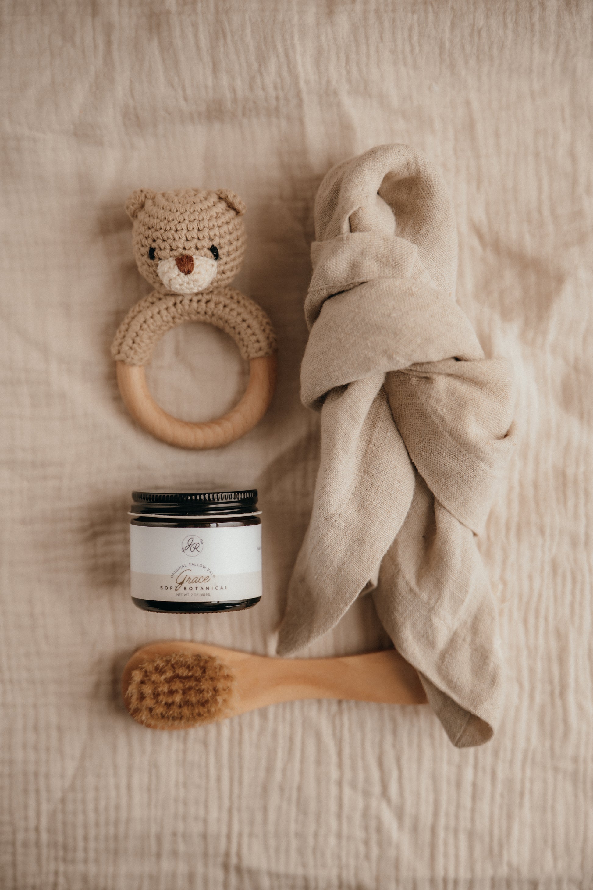 Baby care items including a wooden ring, jar, towel, and brush on a soft surface.