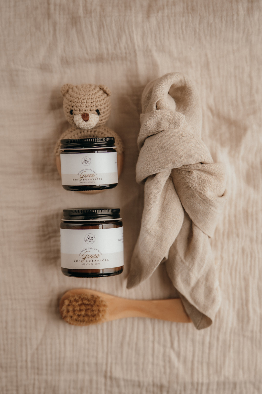 Two jars, a small crocheted bear, a wooden brush, and a beige towel on a textured surface.