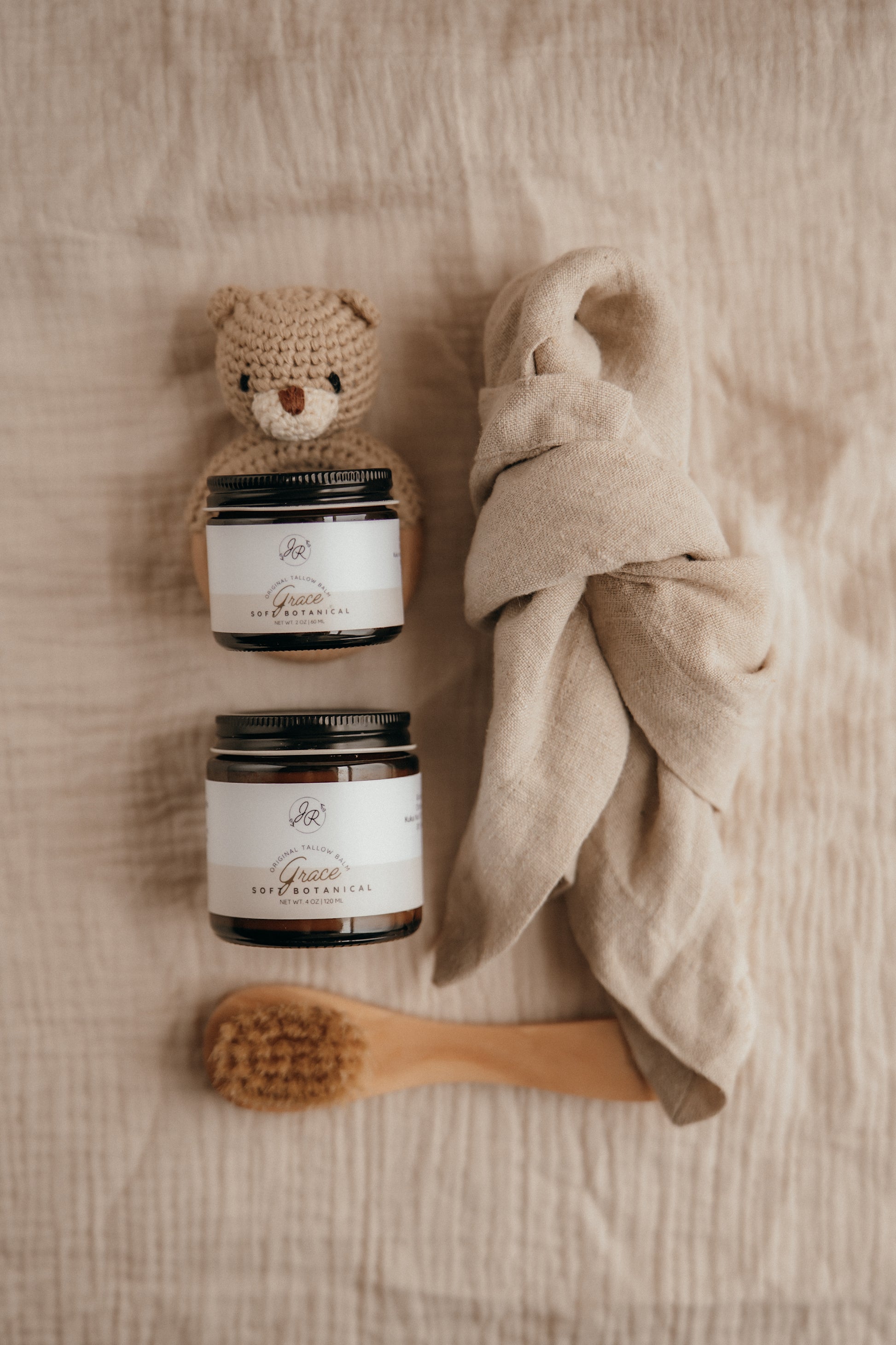 Two jars, a small crocheted bear, a wooden brush, and a beige towel on a textured surface.