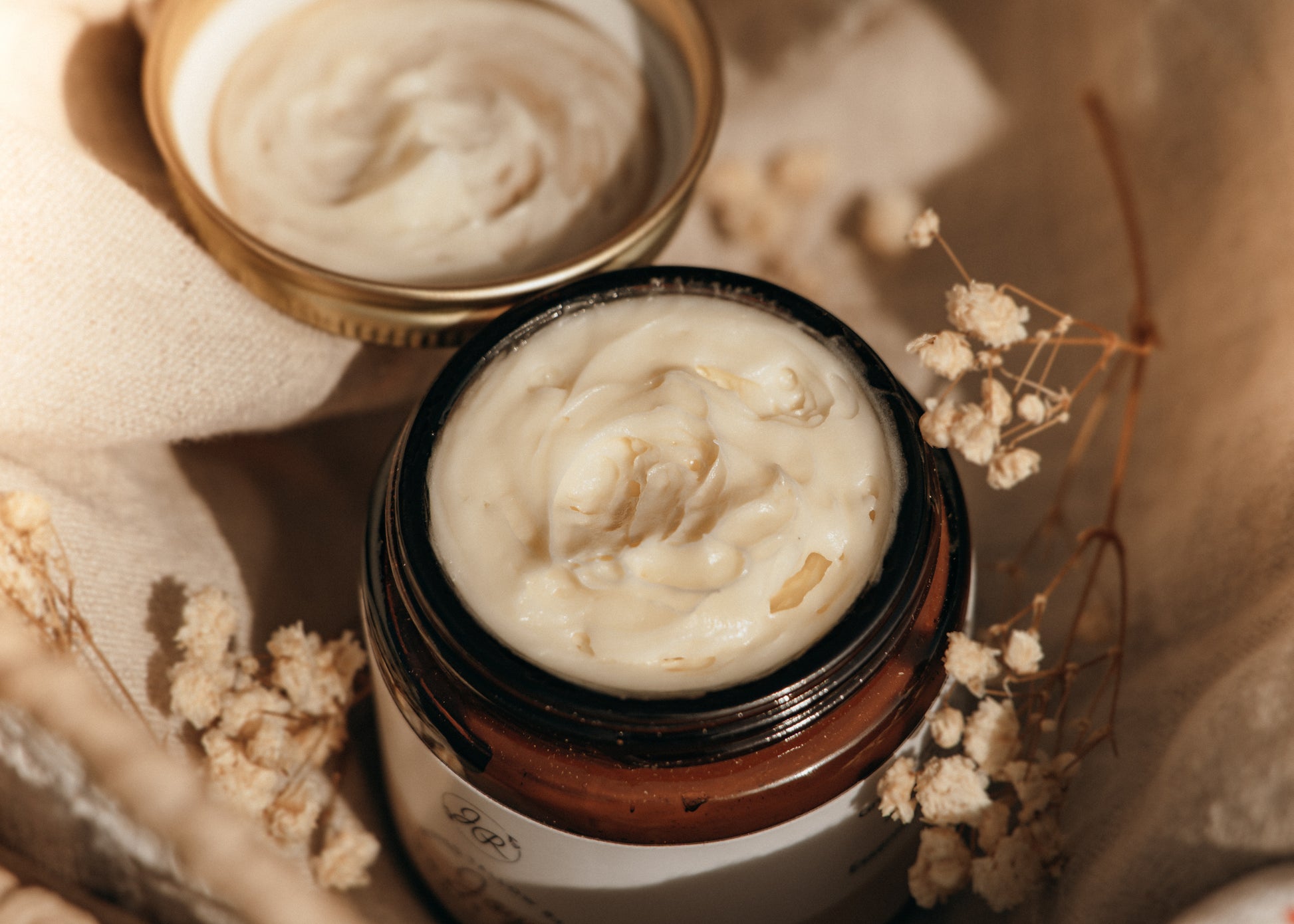 Jar of cream with a lid on a textured surface with dried flowers.