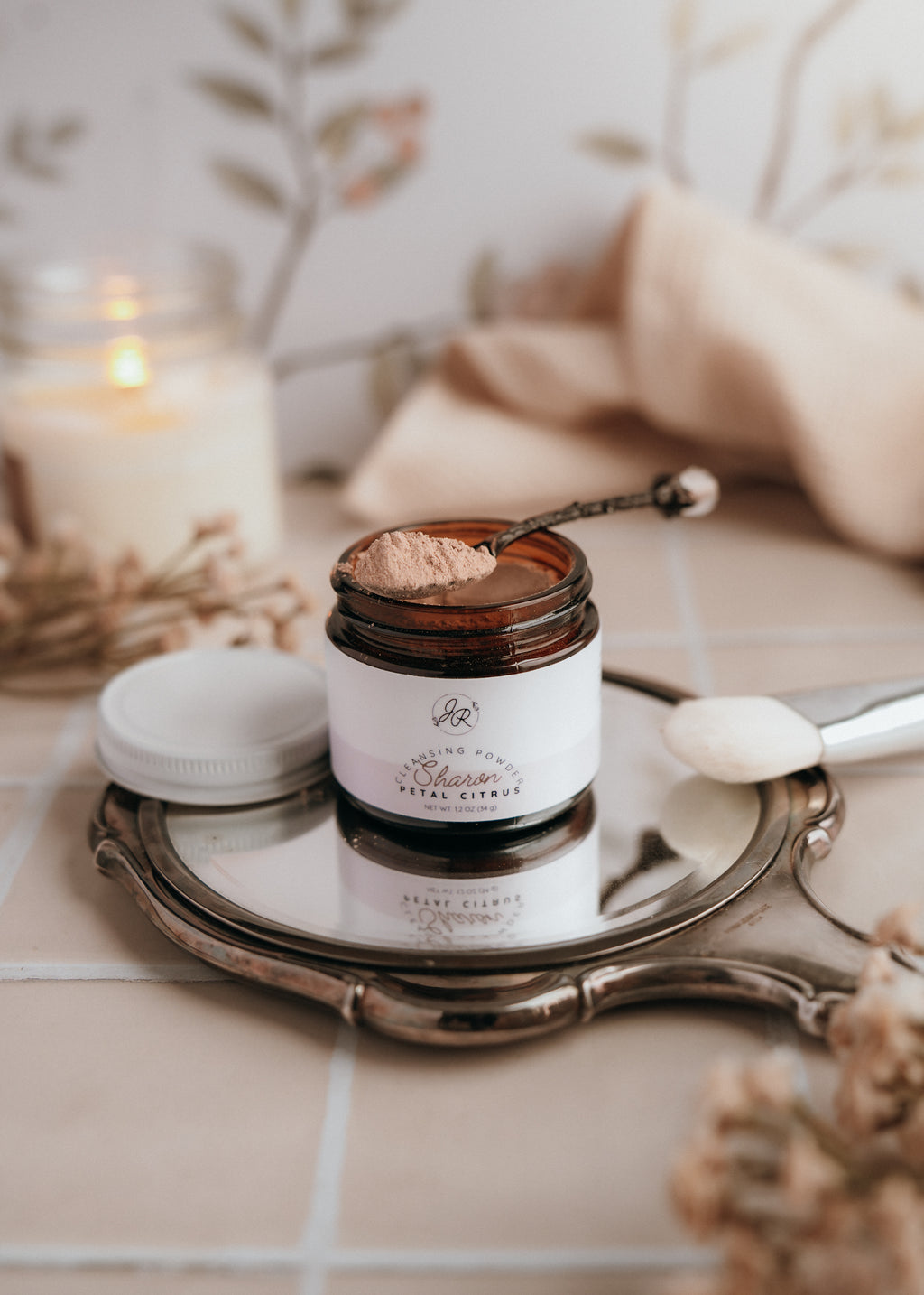 Skincare product jar with applicator on a decorative tray with candles and flowers in the background